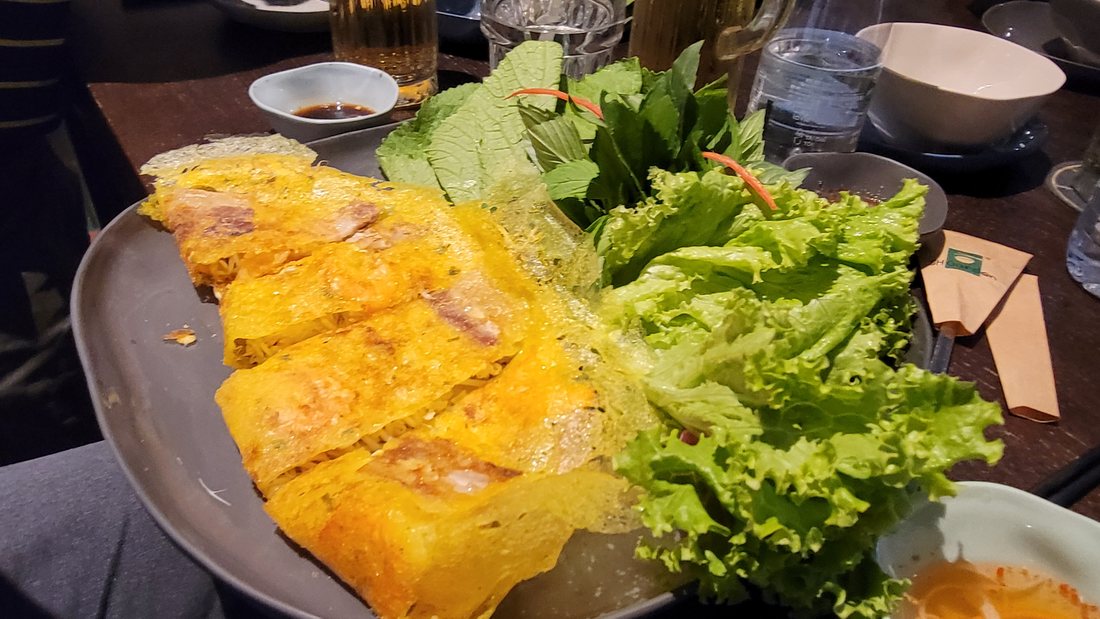 Bánh xèo, folded and cut into strips — the turmeric-yellow crepe, pork and shrimp visible through it, and the herb platter it is meant to be eaten with