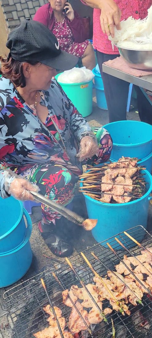 Her at the grill: long tongs in a plastic-gloved hand, a piece of pork lifted off the fire, smoke coming up through two rows of skewers, and a blue bucket of already-grilled ones at her knee. Behind her, someone is lifting a mound of bún out of a steel bowl